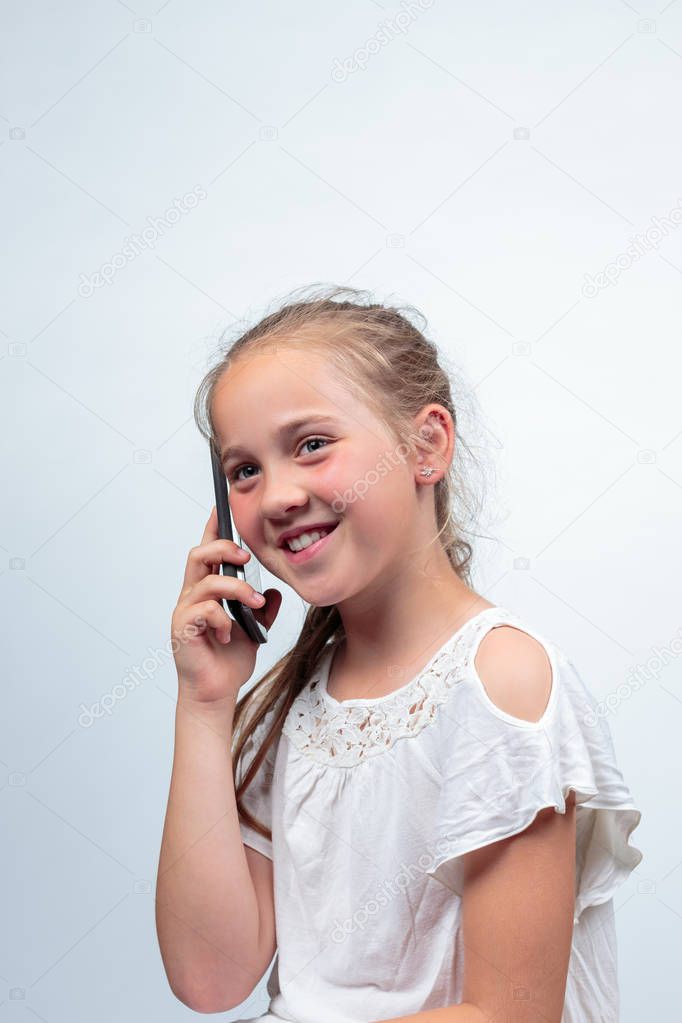 A portrait of a pretty young caucasian girl (10 years old) smiling wearing a white summer dress or blouse making a phone call using a cellphone looking away from camera on a light background