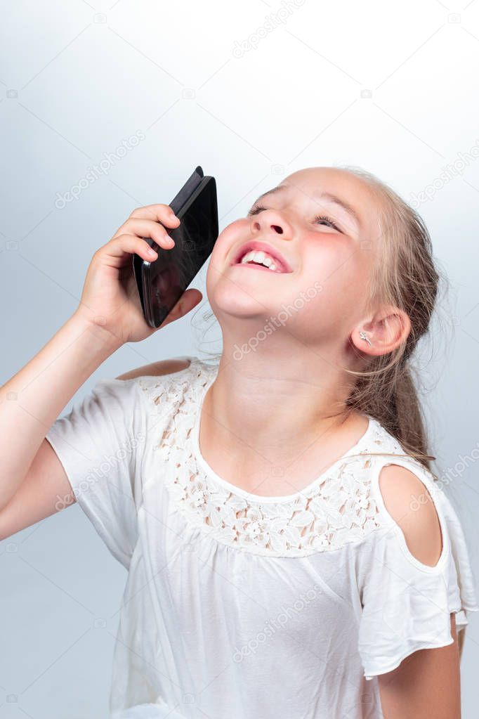 A pretty young caucasian girl (10 years old) in white summer shirt laughing out loud while holding a black mobile phone, positive feelings on a light background