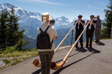 Schynige Platte, Bernese Oberland, İsviçre - 12019 Ağustos: 2 Alpenhorn oyuncuları geleneksel İsviçreli kadın turist tarafından cep telefonuyla filme alındı. Alp panorama arkaplanı
