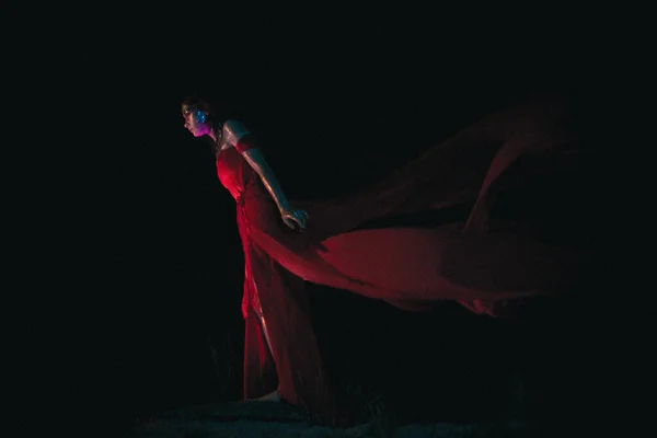 Cinematic goddess stands in the dark, her red dress developing on the wind