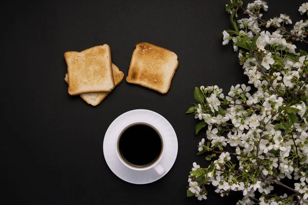 White cup with coffee and Toast, Spring Flowers, Cherry branch on a dark background. Flat lay, top view.