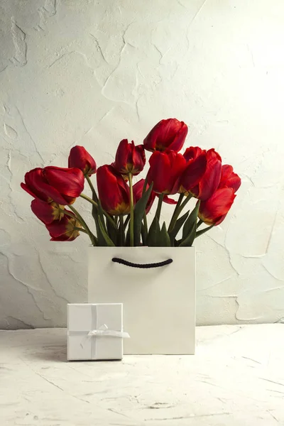 White Gift Bag, small white gift box, petals and bouquet of red tulips on a light stone background. Concept Offer a gift or an engagement, marriage.
