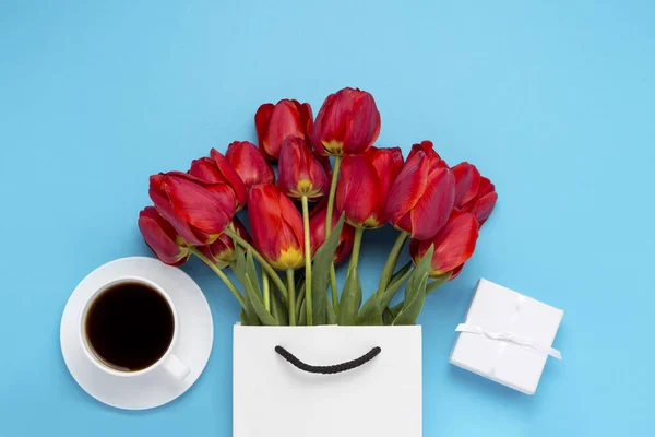White Gift Bag, a small white gift box, a white cup with black coffee and a bouquet of red tulips on a blue background. Concept Offers an engagement or marriage, shopping.