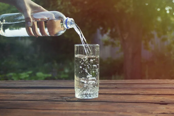 Female hand pours water from a plastic bottle into a glass on a wooden table in a spring garden.