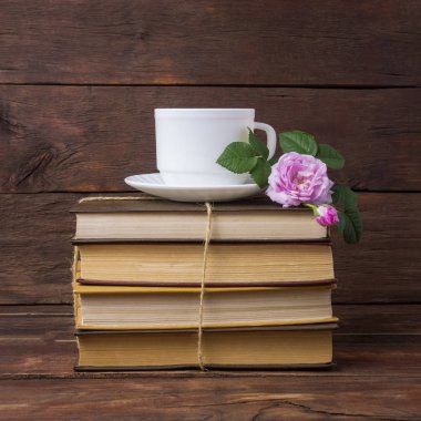 Kitaplar, beyaz fincan kahve ve koyu ahşap arka plan üzerinde pembe bir gül yığını.