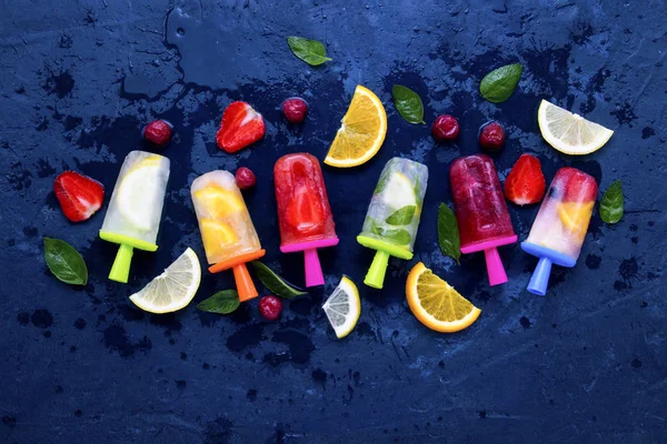 Homemade bright fruit popsicle with strawberry, cherry, lemon, orange, lemon and mint flavor and fresh fruit for ice cream on a dark blue background. Flat lay, top view.