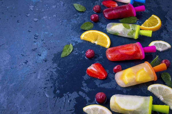 Multicolored bright fruit popsicle with strawberry, cherry, lemon, orange, lemon and mint aroma and fresh fruit on a dark blue background.