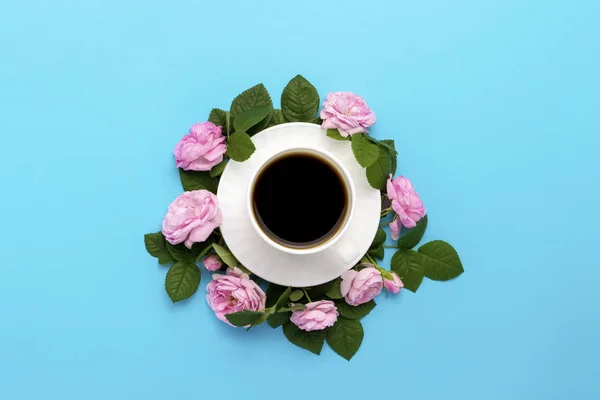 White cup with black coffee and pink roses lined around on a blue background. Flat lay, top view.