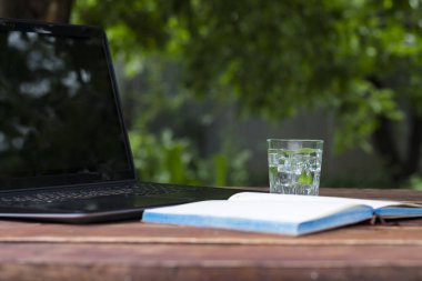Laptop, açık günlüğü ve bir bardak buz küpleri ve nane ile ferahlatıcı bir içki ile ahşap bir masaya bırakır. Doğada, freelancing, tatilde iş iş kavramı.