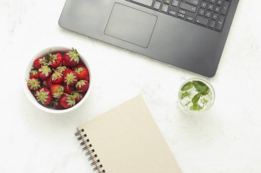 Çalışma alanı. Laptop, defter, beyaz bir kase çilek ve serinletici bir içecek buz ve nane ile. Üstten görünüm, düz yatıyordu