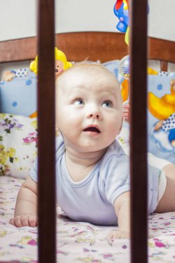 Çok güzel bir yüz ve mavi gözlü küçük bebek bebek karyolası/beşik midesinde üzerinde yatıyor ve çocuklar için mavi bir koruma ile baktı. Resmi yatağın parmaklıkların arasından alınır.