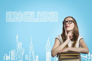 Gözlüklü genç bir kız kitap yığınına yaslanıyor ve mavi arka planda dalgın bir yüzle İngilizce metniyle yukarı bakıyor. İngiltere ve ABD 'de İngilizce ve eğitim kavramı.