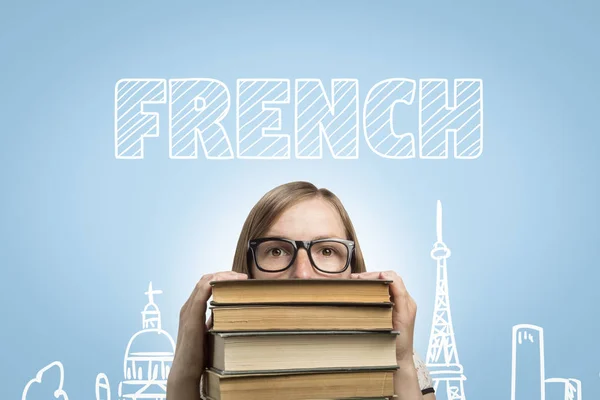 Young girl student with glasses peeks out from under a pile of books on a blue background with text French. Concept of learning French and education in France.