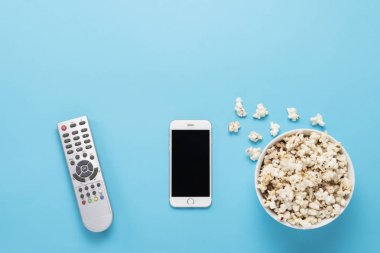 Patlamış mısırlı kase, uzaktan kumandalı televizyon, mavi arka planda beyaz telefon. Ev sineması, sinema, boş zaman kavramı. Düz yatış, üst görünüm.