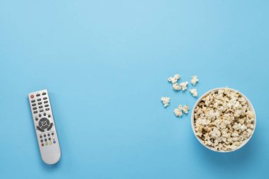 Mavi arka planda patlamış mısır ve uzaktan kumandalı televizyon kasesi. Ev sineması, sinema, boş zaman kavramı. Düz yatış, üst görünüm.
