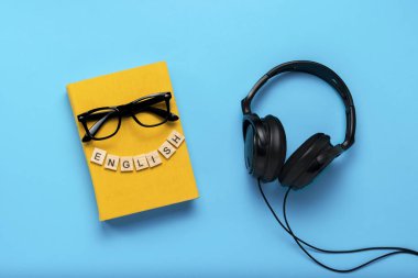 Book with a yellow cover with text English, glasses and black headphones on a blue background. Concept of audio books, self-education and learning English independently. Flat lay, top view. clipart
