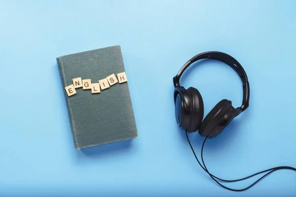 Book with a blue cover with text English and black headphones on a blue background. Concept of audio books, self-education and learning English independently. Flat lay, top view.