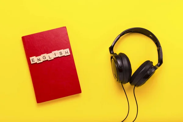 Book with a red cover with text English and black headphones on a yellow background. Concept of audio books, self-education and learning English independently. Flat lay, top view.