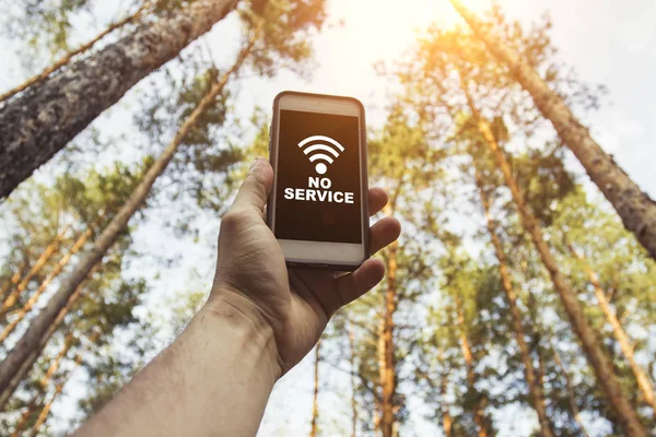 Male hand is holding a phone with sign no service  above his head against the background of trees in a pine forest. Concept of no service, wifi, internet in the forest or a place far from the city.