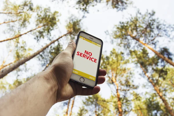 Male hand is holding a phone with sign no service  above his head against the background of trees in a pine forest. Concept of no service, wifi, internet in the forest or a place far from the city.