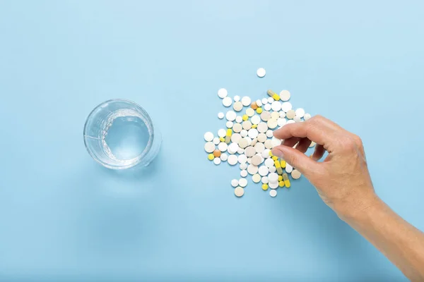 Female hand takes a pill from many others, a glass of water on a blue background. Concept of taking drugs in case of illness and disease. Flat lay, top view.