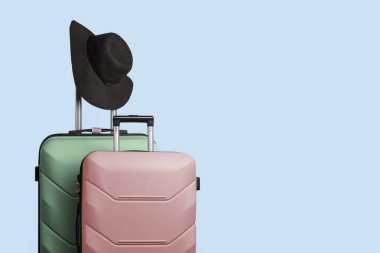 Two plastic suitcases on wheels and a wide-brimmed hat on the handle of a large suitcase against a blue background. Travel concept, vacation trip, visit to relatives. clipart
