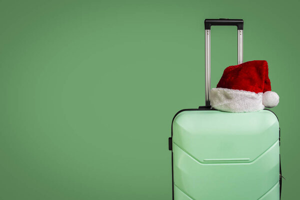 Plastic suitcase and Santa Claus hat on green background. Concept of travel to visit friends and relatives on Christmas holidays. Merry Christmas and Happy New Year. Christmas trip.