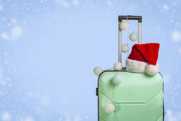 Plastic suitcase, Santa Claus cap and garland on a blue background with snow. Concept of travel, business trips, trips to visit friends and relatives on Christmas holidays. New Year's journey.