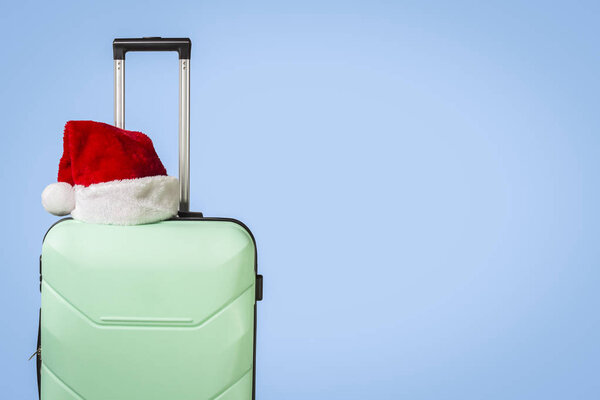 Plastic suitcase and Santa Claus hat on blue background. Concept of travel to visit friends and relatives on Christmas holidays. Merry Christmas and Happy New Year. Christmas trip.