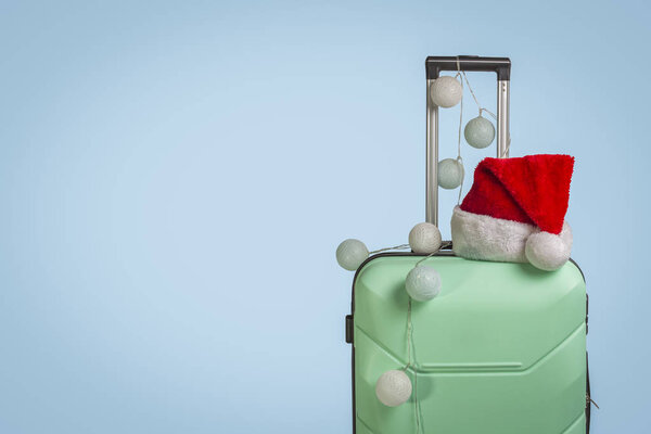 Plastic suitcase, Santa Claus cap and garland on a blue background. Concept of travel, business trips, trips to visit friends and relatives on Christmas holidays. New Year's journey.