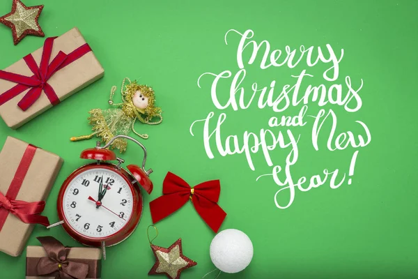 Red alarm clock, Christmas decorations, Christmas toys and gift boxes on a green background. Added text Merry Christmas and Happy New Year. Flat lay, top view.