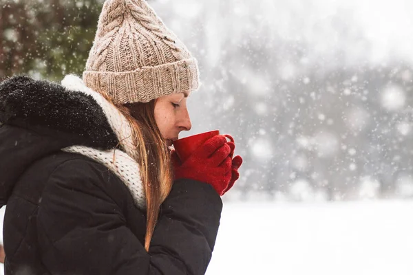 Young Girl in Red Gloves holds a Red Cup with Hot Tea on the Background of the Winter Landscape during Snowfall.