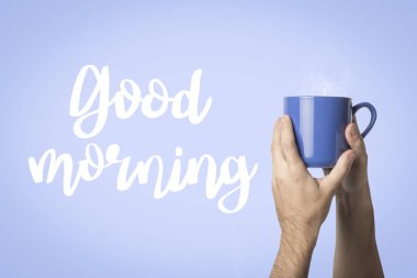 Erkek elini mavi bir fincan sıcak kahve veya çay mavi bir arka plan üzerinde tutuyor. Metin Günaydın ekledi. Sıcak kahve ile kahvaltı konsepti.