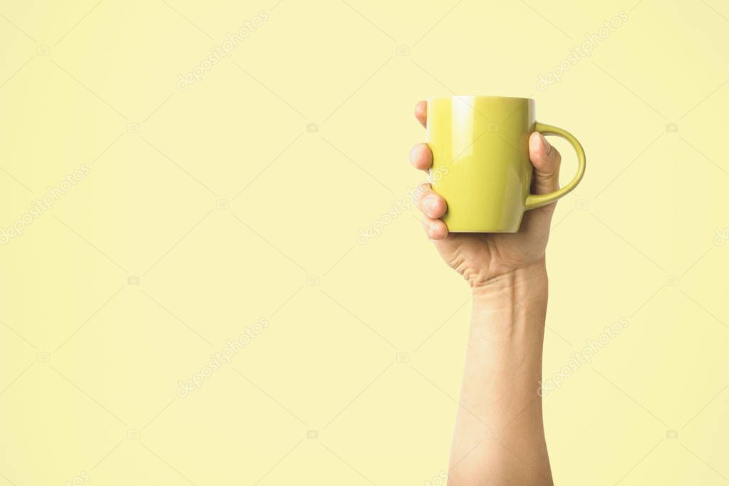 Female hand holding a green cup with hot coffee or tea on a light yellow background. Breakfast concept with hot coffee or tea.