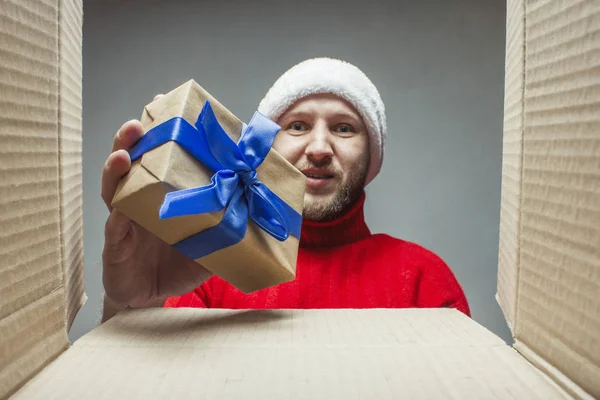 Man in a Santa Claus hat and a red sweater lays down, takes gifts in or out of the box. Concept of packaging and sending or receiving gifts for friends and relatives for Christmas, New Year
