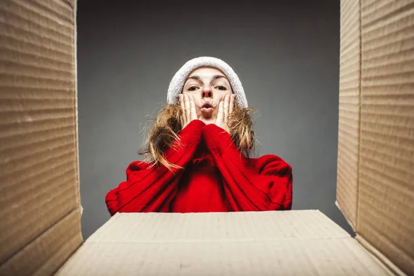 Girl with a surprised face in a Santa Claus hat and a red sweater looks at the contents of the box. Concept of opening gifts, surprise, gift from friends, relatives, parcel, shopping