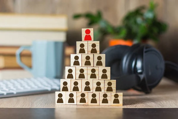 Wooden cubes with men lined up with a pyramid on the background of an office desk with a laptop, keyboard, headphones, mug. Concept of corporation, financial pyramid, leadership, united team