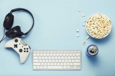 Keyboard and tin can with a drink, energy drink, a bowl of popcorn, a gamepad and headphones on a blue background. The concept of computer games, entertainment, gaming, leisure. Flat lay, top view clipart
