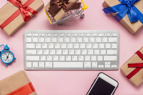Keyboard, alarm clock, mobile phone and gift boxes laid out around the keyboard on a pink background. Concept of ordering and buying gifts on the Internet, online store. Flat lay, top view