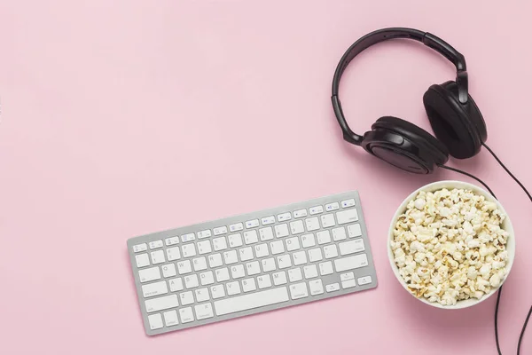 Keyboard, headphones and a bowl of popcorn on a pink background. The concept of watching movies, shows, sports on the PS, games online. Flat lay, top view