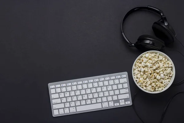 Keyboard, bowl with popcorn and headphones on a black background. The concept of the game on the PC, gaming, watching movies, TV shows, sports competitions on the PC. Flat lay, top view