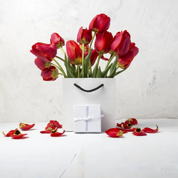 White Gift Bag, small white gift box, petals and bouquet of red tulips on a light stone background. The concept of an offer for an engagement or a marriage.