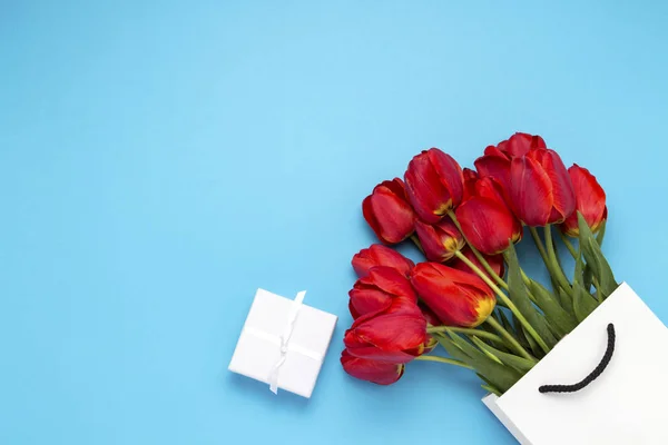 White Gift Bag, a small white gift box and a bouquet of red tulips on a blue background. Concept Offers an engagement or marriage, shopping.