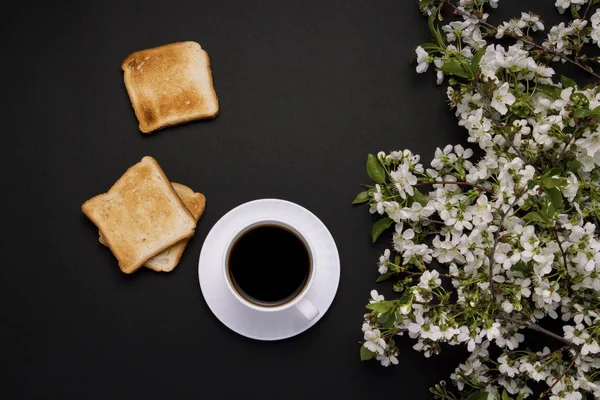 White cup with coffee and Toast, Spring Flowers, Cherry branch on a dark background. Flat lay, top view.