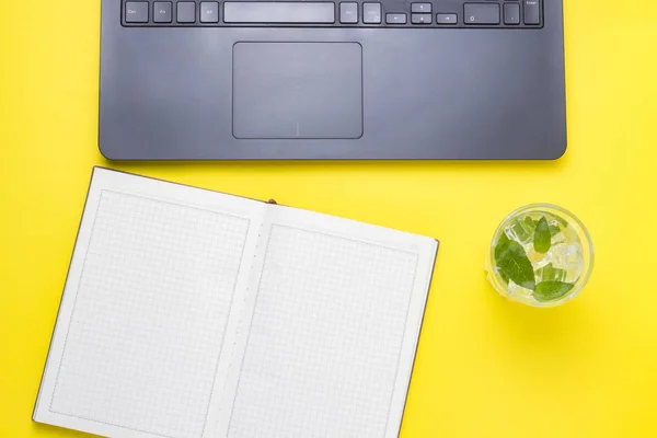 Workplace with Laptop, a fresh drink with ice and mint, an open diary on a yellow background. The concept of the workspace of a journalist, writer, freelancer, copywriter. Flat lay, top view.