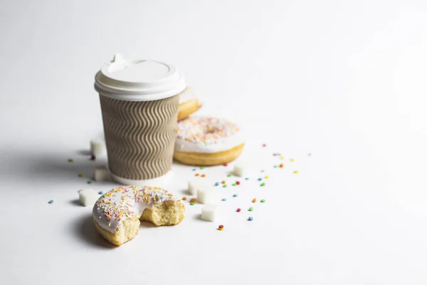 A paper cup with a lid, coffee or tea to go and fresh tasty donuts and sweet multicolored decorative candies on a white background. Bakery concept, fresh pastries, delicious breakfast, fast food