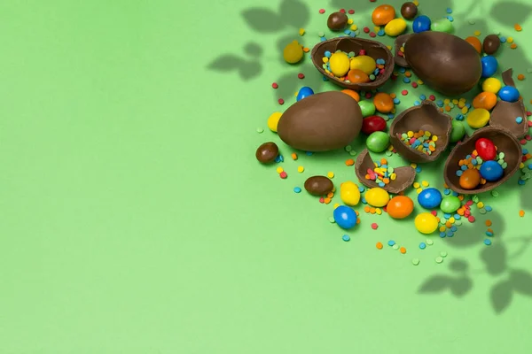 Broken and whole chocolate Easter eggs, multicolored sweets on a green background. Shrub. Concept of celebrating Easter, Easter decorations. Flat lay, top view. Copy Space.