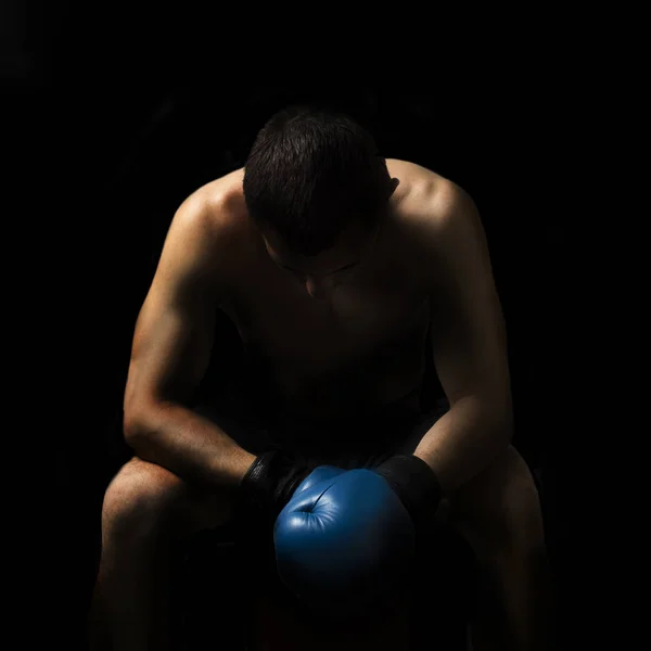 A man in boxing gloves is sitting on a chair in the corner of the ring against a dark background. Boxing concept, boxing training, sparring
