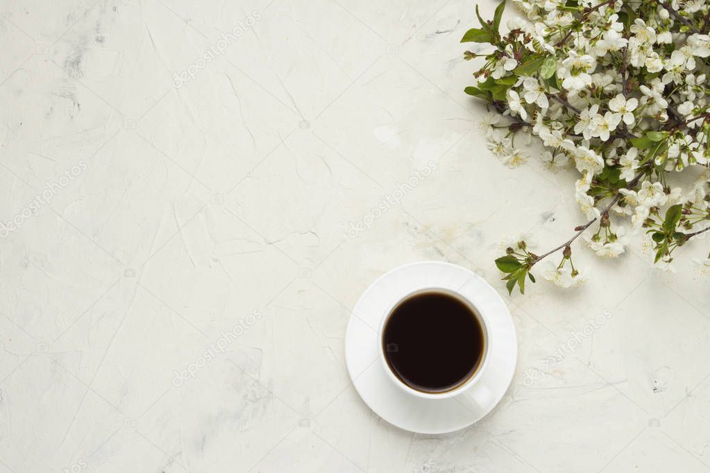 cup with black coffee, branches with white flowers on a light stone background. Flat lay, top view