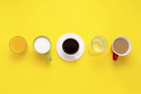 Grupo de bebidas en tazas multicolores, café negro, café con leche, yogur, agua, jugo de naranja se ponen en una línea sobre un fondo amarillo. Piso tendido, vista superior — Foto de Stock